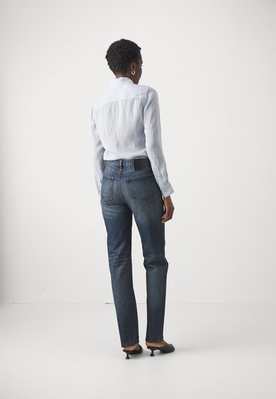 Lichtblauw, gestructureerd shirt met een button-down kraag, gecombineerd met donkere denim jeans met twee achterzakken, gedragen met zwarte hakken.