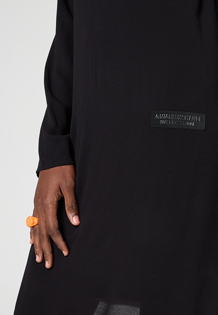 Indumento nero con una texture liscia, dotato di un'etichetta nera con logo. Mano mostrata mentre indossa un anello rotondo arancione.