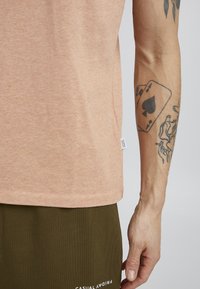 Tricou scurt din piersică deschisă, cu mânecă scurtă și guler rotund, realizat dintr-un material moale, având un etichetă pe lateral și o pereche de pantaloni de culoare verde măslin cu text alb.