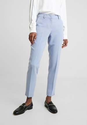 Personne portant un pantalon slim bleu clair, une chemise blanche à manches longues et des mocassins noirs se tenant sur fond blanc uni.