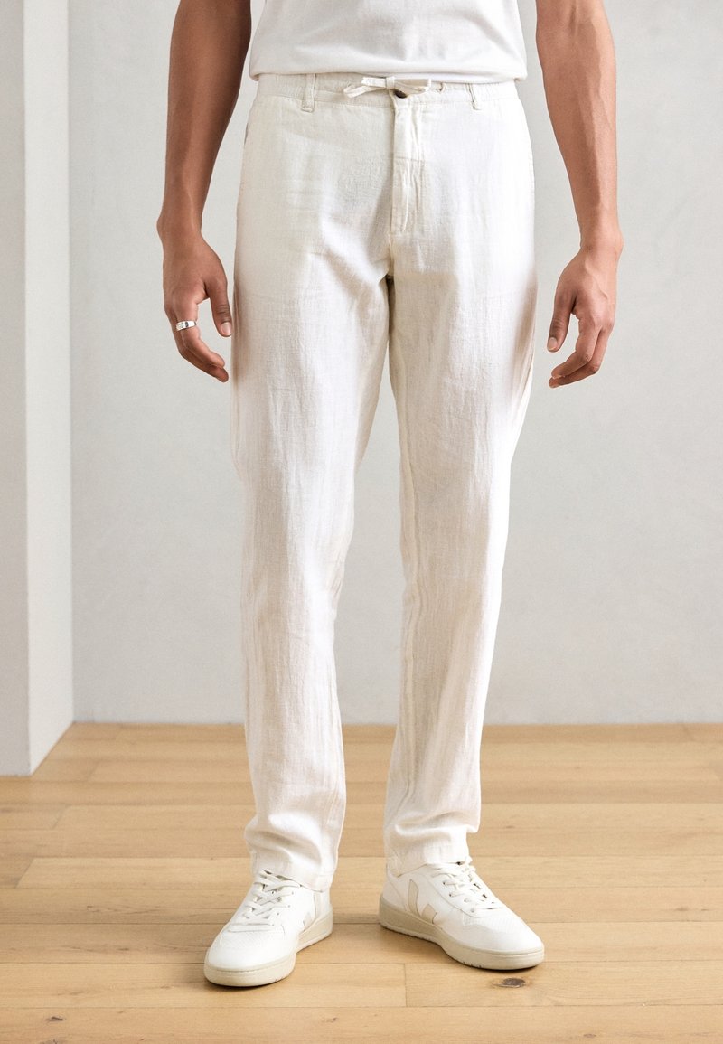 Pantalons en lin blancs avec une taille élastique à cordon et des poches latérales, associés à des baskets blanches avec des accents beiges subtils.