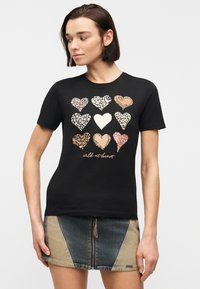 Γυναίκα που φοράει μαύρο T-shirt με εννέα καρδιές σε λεοπάρ σχέδιο και το κείμενο «wild at heart», σε συνδυασμό με δίχρωμη τζιν φούστα με φερμουάρ μπροστά.