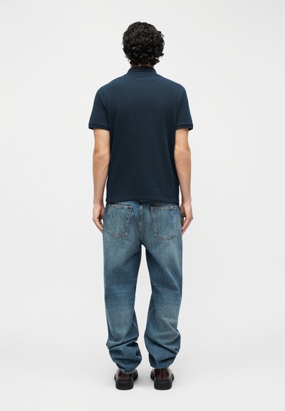 Άνδρας με μπούκλες, με την πλάτη γυρισμένη, φοράει μπλε μπλουζάκι πόλο, χαλαρά μπλε τζιν και σκούρα καφέ παπούτσια, σε απλό φωτεινό φόντο.