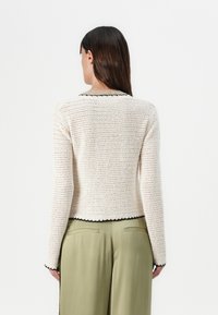 Cardigan a maglia di colore bianco sporco con dettagli neri; design accorciato, maniche lunghe, tessuto texturizzato e orlo scallop. Indossato con pantaloni verdi.