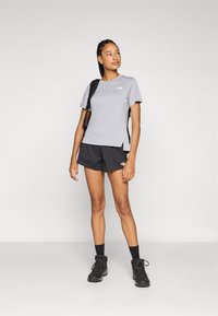 Grijze T-shirt met korte mouwen en logo, gecombineerd met zwarte shorts met een gestructureerd patroon. Het model draagt wandelschoenen en een rugzak.