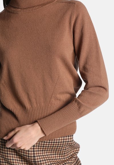 Pull à col roulé côtelé marron porté par une personne avec la main droite posée sur une jupe ou un pantalon à motif pied-de-poule.