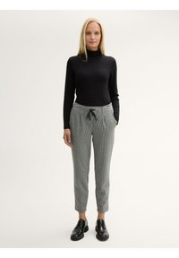 Schwarzer gerippter Rollkragenpullover und graue karierte Hose mit Kordelzug, Fronttaschen und schmal zulaufendem Bein. Schwarze Schuhe runden das Outfit ab.