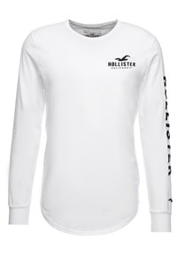 Vit långärmad t-shirt i bomull, med en liten svart logotyp på bröstet och svart text längs den vänstra ärmen.