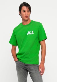 Vyras vilkintis ryškiai žalią Calvin Klein marškinėlį su "ALL" logotipu, pozuoja su viena ranka kišenėje ir žiūri į šoną prieš baltą foną.