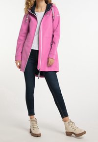 Ljusrosa lång jacka med dragkedja fram, marinblå huvligning, två sidofickor och vita kontrasterande detaljer. Bärs med mörka jeans och beiga stövlar.