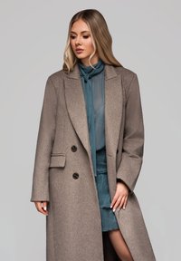 Lang, skræddersyet frakke i lys brun uldblanding med to knapper og sidelommer. Båret over en teal gennemsigtig bluse og en struktureret teal nederdel.