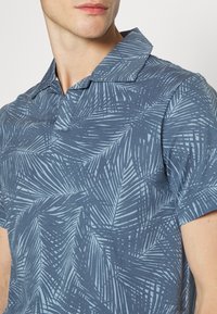 Polo à manches courtes bleu affichant un motif de feuilles de palmier bleu clair, fabriqué en tissu doux avec un col pointu et une patte de boutonnage à trois boutons.