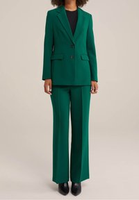 Grön skräddarsydd kostym med en strukturerad kavaj och vida byxor. Slät tyg, två knappar och sidofickor framhäver designen.