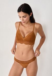 Woman with dark hair wearing a matching brown lace bra and panties set, adjusting her panties, standing against a plain light background.