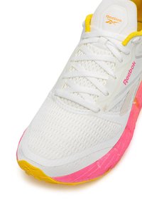 Fehér sportcipő texturált felsőrésszel, sárga kiegészítőkkel és rózsaszín gumi talppal. Fehér fűzőkkel és Reebok logóval a nyelvén.