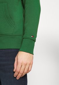 Ręka oparta luźno obok ciała, ubrana w zieloną bluzę z małym czerwono-biało-czarnym logo na mankiecie rękawa oraz ciemnoniebieskie jeansy.