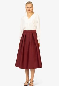 Blusa bianca a maniche lunghe con scollatura a V abbinata a una gonna midi plissettata bordeaux, con una texture liscia e una vita ampia. Décolleté neri con cinturini.