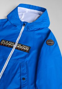 Blå huva jacka gjord av lätt material, med en vit nätfoder, logopatch och dragkedja fram med knäppfickor.
