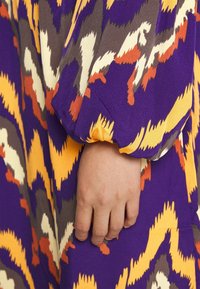 Main reposant contre un tissu avec un motif abstrait en zigzag violet, jaune, blanc, orange et gris, et un poignet froncé sur la manche.