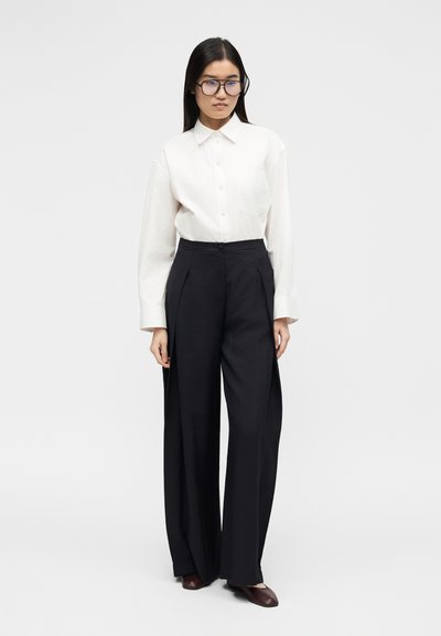 Γυναίκα με μακριά σκουρόχρωμα μαλλιά και γυαλιά, φοράει ένα λευκό πουκάμισο με κουμπιά, μαύρο παντελόνι φαρδύ και σκούρα καφέ παπούτσια, στέκεται σε απλό φόντο.