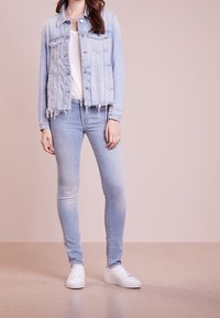 Světle modrá denimová bunda s třásněmi na lemu, nošená přes bílou tričko, spárovaná se světle modrými skinny džíny a bílými teniskami.