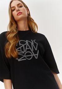 Czarna, oversize koszulka z białym graficznym napisem. Materiał wydaje się miękki, o gładkiej teksturze i krótkich rękawach.