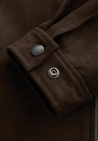 Brown suede jacket close-up featuring a buttoned cuff with black and gold accents, highlighting soft texture and detailed stitching.