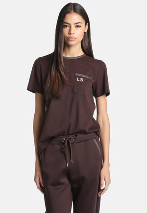 Jeune femme portant un t-shirt marron foncé à manches courtes avec "LS" sur la poche et un pantalon à cordon assorti, debout devant un fond uni.