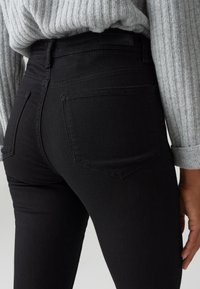 Personne portant un jean noir ajusté et un pull gris clair côtelé avec les manches retroussées, vue de dos de la taille aux pieds.