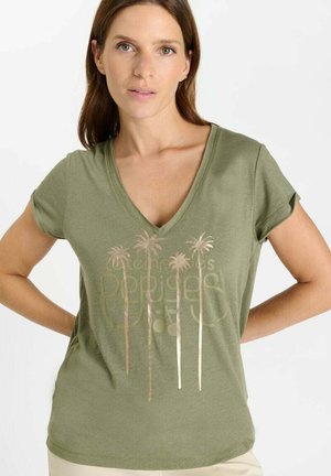 Mujer con camiseta de cuello en V color verde oliva con diseños metálicos de palmeras y texto tenue, de pie con las manos en las caderas contra un fondo blanco.