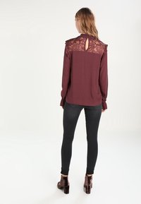 Blusa bordeaux a maniche lunghe con dettagli in pizzo sulle spalle, scollatura a chiave sul retro, abbinata a jeans skinny neri e stivali con tacco.