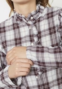 Kvinde justerer manchetterne på en langærmet, lilla og hvid ternet bluse med flæsekrave og knaplukning foran.