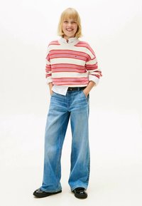 Giovane sorridente con capelli biondi che indossa un maglione a righe rosa e bianco, jeans a gamba larga blu e scarpe nere, in piedi con le mani in tasca.