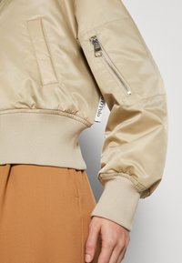 Veste bomber beige avec poignets et ourlet côtelés, poche zippée sur la manche, portée avec un pantalon marron, montrant partiellement le bras et la main.