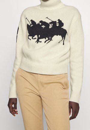 Pull à col roulé crème avec silhouette noire de trois joueurs de polo à cheval, porté avec un pantalon beige sur une personne à la peau foncée.
