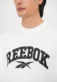 Bijela majica s okruglim izrezom s velikim crnim logotipom "REEBOK" koji ima karakterističan zakrivljeni naglasak ispod. Tkanina izgleda glatko i lagano.