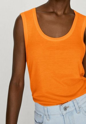 Mørkhudet person iført en knaldorange ærmeløs ribbet top, der er stoppet ned i lyseblå højtaljede jeans med knaplukning.