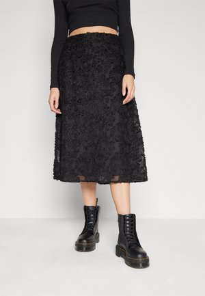 Personne portant une jupe midi noire texturée, un top court à manches longues noir et des bottes de combat noires lacées.