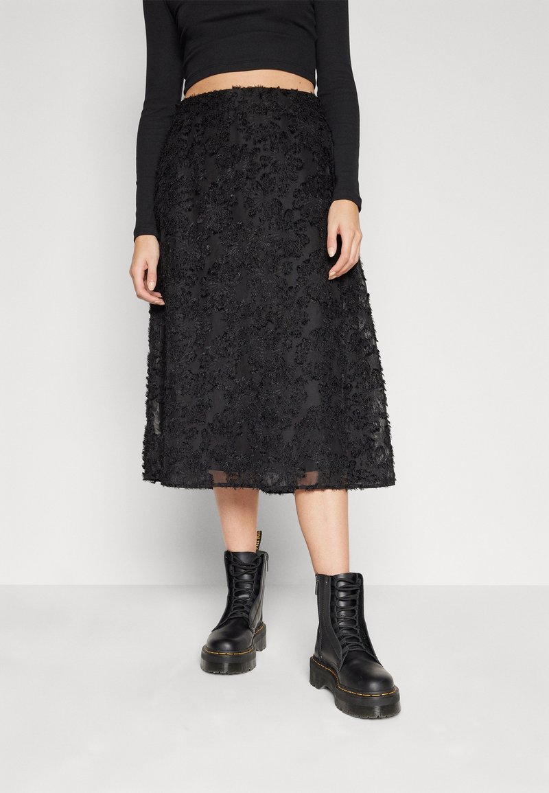 Personne portant une jupe midi noire texturée, un top court à manches longues noir et des bottes de combat noires lacées.