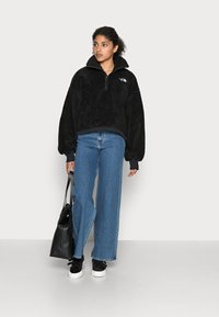 Pullover in pile nero con colletto alto e zip a quarto, abbinato a jeans blu a vita alta. Sneakers nere e borsa a tracolla nera.