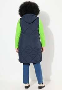 Granatowa pikowana kamizelka puchowa z kapturem, założona na zieloną koszulę z długim rękawem. Posiada kieszenie zapinane na zamek oraz długi krój, zestawiona z niebieskimi dżinsami.