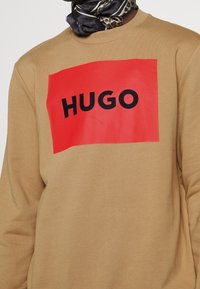 Persona que lleva una sudadera color beige con un gran rectángulo rojo y el texto negro en negrita "HUGO" en el pecho, combinada con una bufanda estampada.