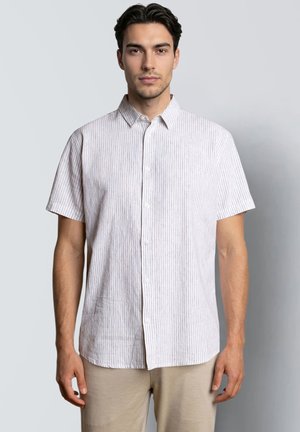 Joven con una camisa blanca de manga corta con botones y finas rayas verticales, de pie frente a un fondo liso de color gris claro.