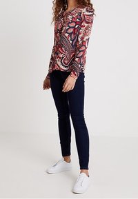 Langärmlige Bluse mit buntem Paisleymuster, strukturiertem Stoff und rundem Ausschnitt, kombiniert mit dunklen Skinny-Jeans und weißen Sneakern.
