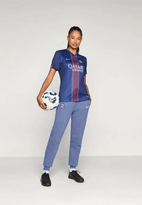 Tumehalli sinine jalgpallipaita punaste detailidega, millel on "Qatar Airways" logo, koos helesiniste pükstega ja mustade jalgpallijalgadega. Hoides valget jalgpall.