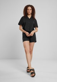 Camicia nera a maniche corte con bottoni abbinata a pantaloni corti coordinati, in tessuto testurizzato. Il modello porta sandali neri con cinturini.