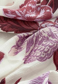 Tissu blanc avec un tissage texturé présentant de grands motifs floraux aux teintes rouge foncé et violet, montrant des plis et des froissements doux.
