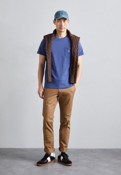 T-shirt de algodão azul com um bolso no peito, colete de malha castanho pousado sobre os ombros, chinos bege e sapatilhas pretas com detalhes brancos.