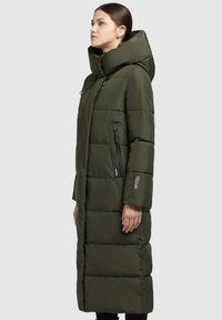 Giacca puffer verde oliva con silhouette lunga, grande cappuccio, tasche con zip e texture trapuntata. Dettagli in hardware neri ovunque.