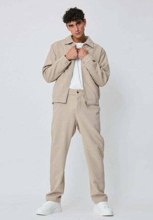 Veste beige zippée avec col, assortie à un pantalon beige. Les deux sont confectionnés dans un tissu doux et texturé. Un t-shirt blanc et des baskets complètent la tenue.